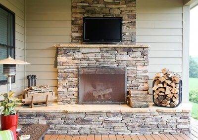 An outdoor wooden fireplace and TV mounted above the fireplace on the stone. Bundles of firewood sit next to the fireplace.