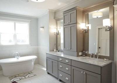 A master bathroom with marble countertops, grey cabinetry, two sinks, and a large whirlpool bathtub below a set of windows.