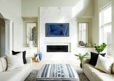 A living room with white couches, gray rug, and houseplants with a marble fireplace and mantle.