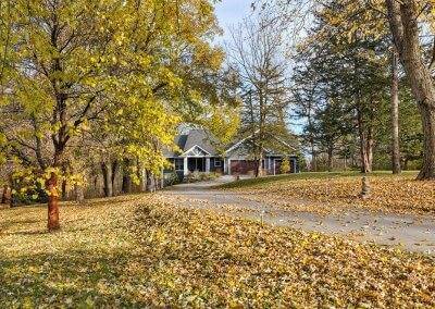 A photo of a house taken from the end of the front yard. Large trees and yellow leaves cover the ground and the house sits nested back in the trees.