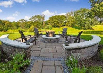 Circular shaped fire pit with Adirondack chairs, sitting wall and lighting.